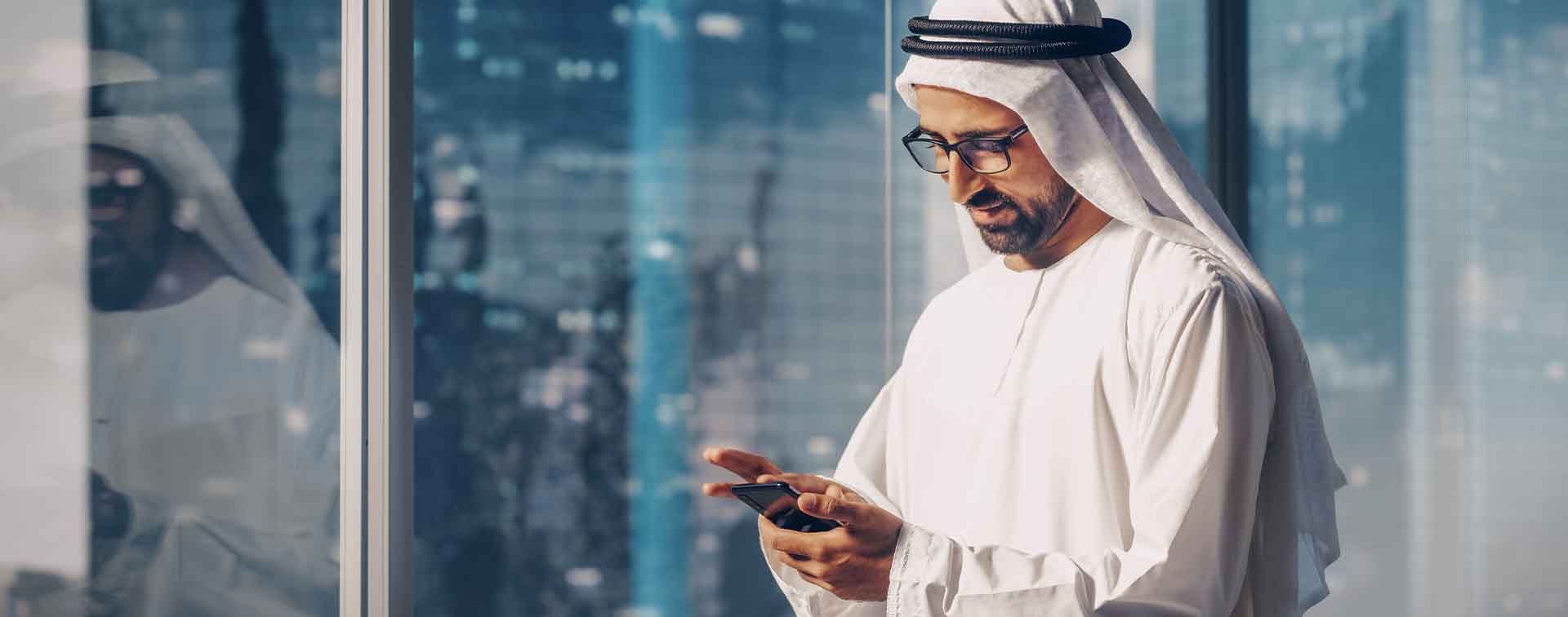 Middle Eastern businessman in traditional outfit in front of a window typing on his cellphone