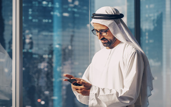 Middle Eastern businessman in traditional outfit in front of a window typing on his cellphone