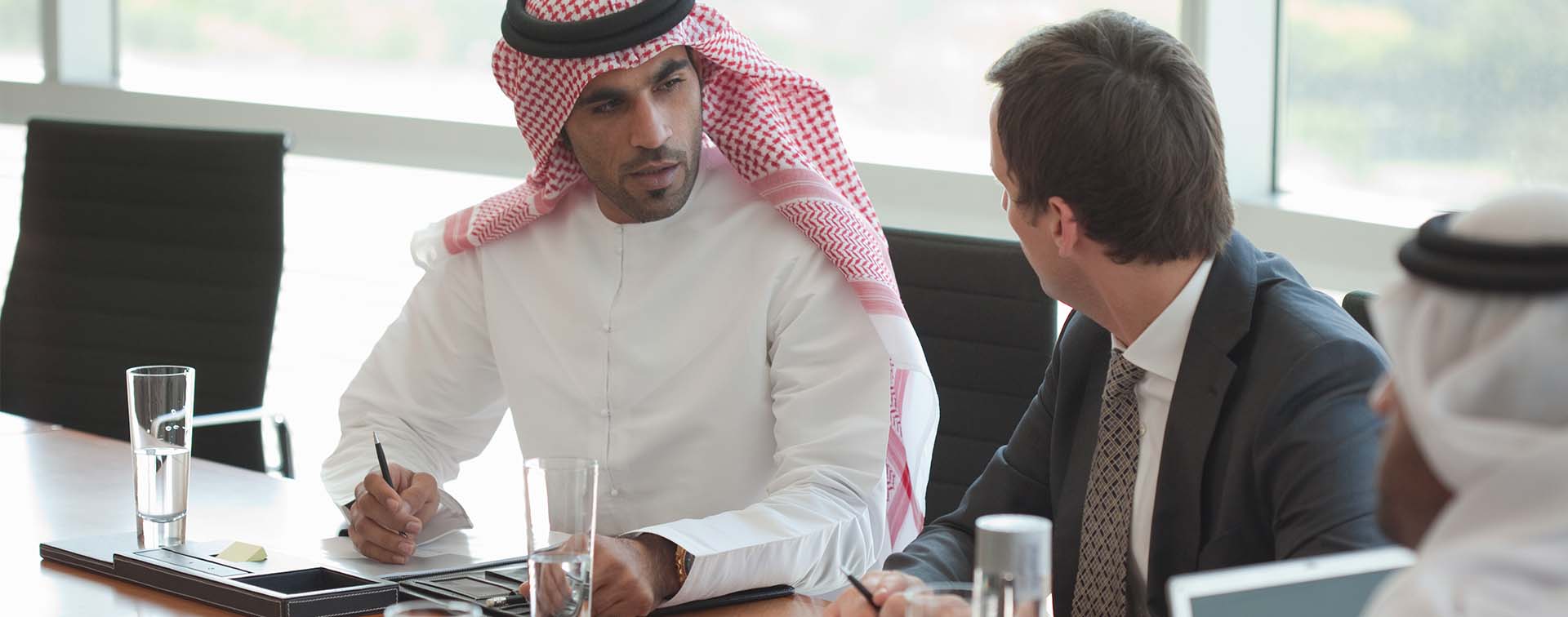 Three businessmen in discussion around a boardroom table, two in Arabic attire and one in a traditional suit