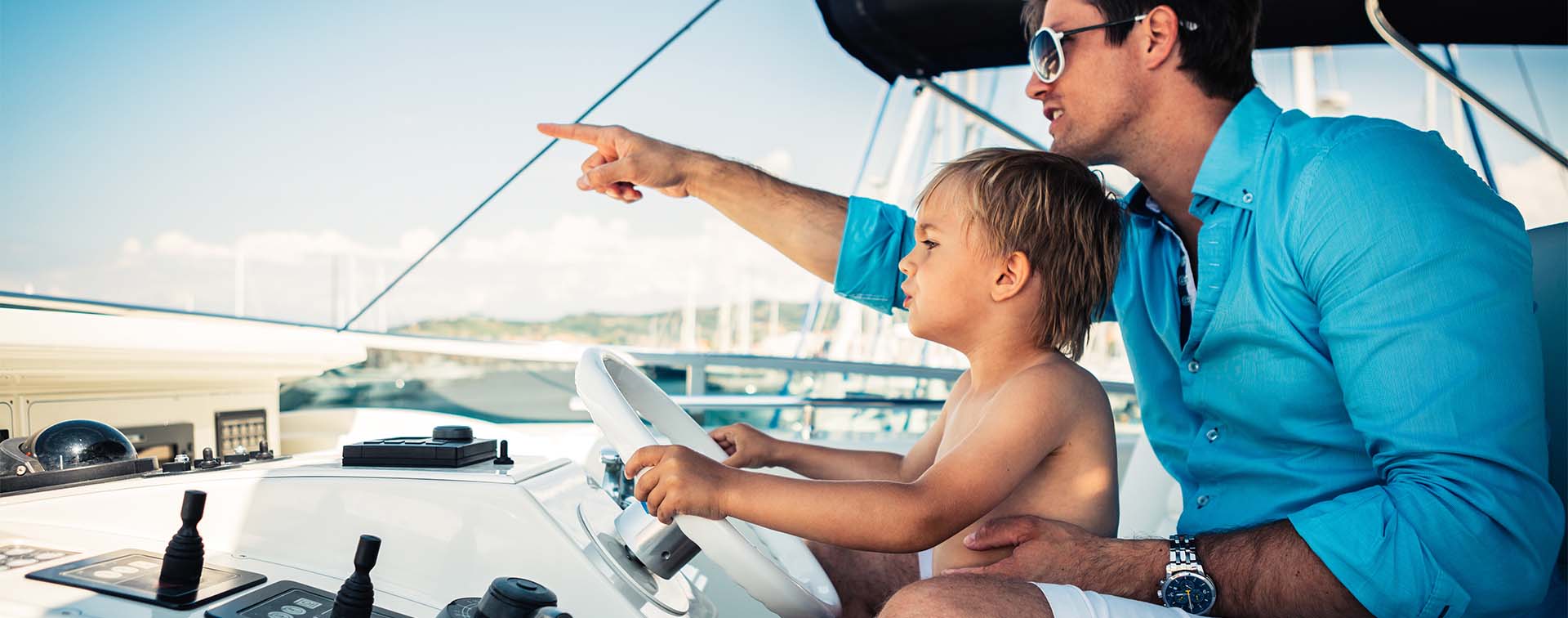 A man on a boat pointing at the distance, with his young son at his lap and holding the steering wheel