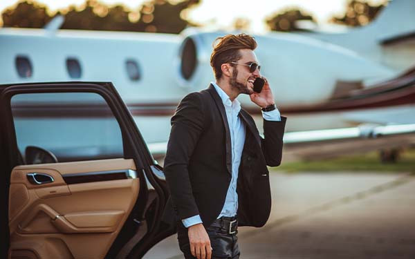 Smiling affluent man talking on the phone while getting out of a car with a private jet in the background