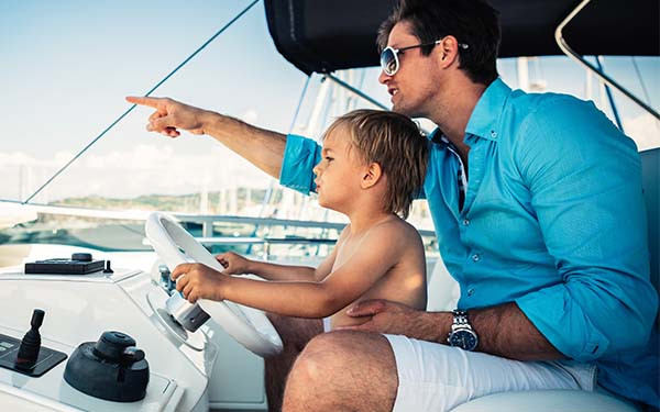 A man on a boat pointing at the distance, with his young son at his lap and holding the steering wheel