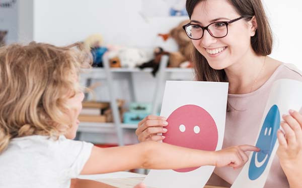A child sitting across their teacher points to one of two smiley face images that she is holding up