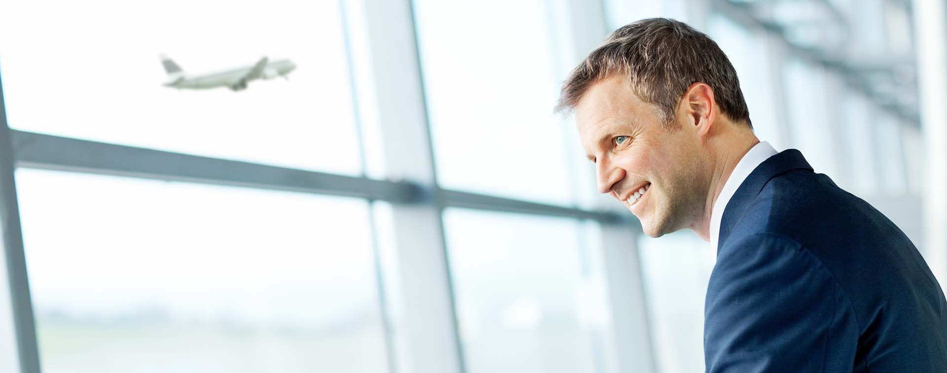 Smiling businessman looking to the left in front of large glass airport window as a plane takes off from the runway in the background