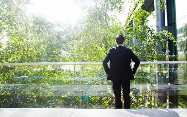 Businessman on a glass balcony looking at trees contemplatively