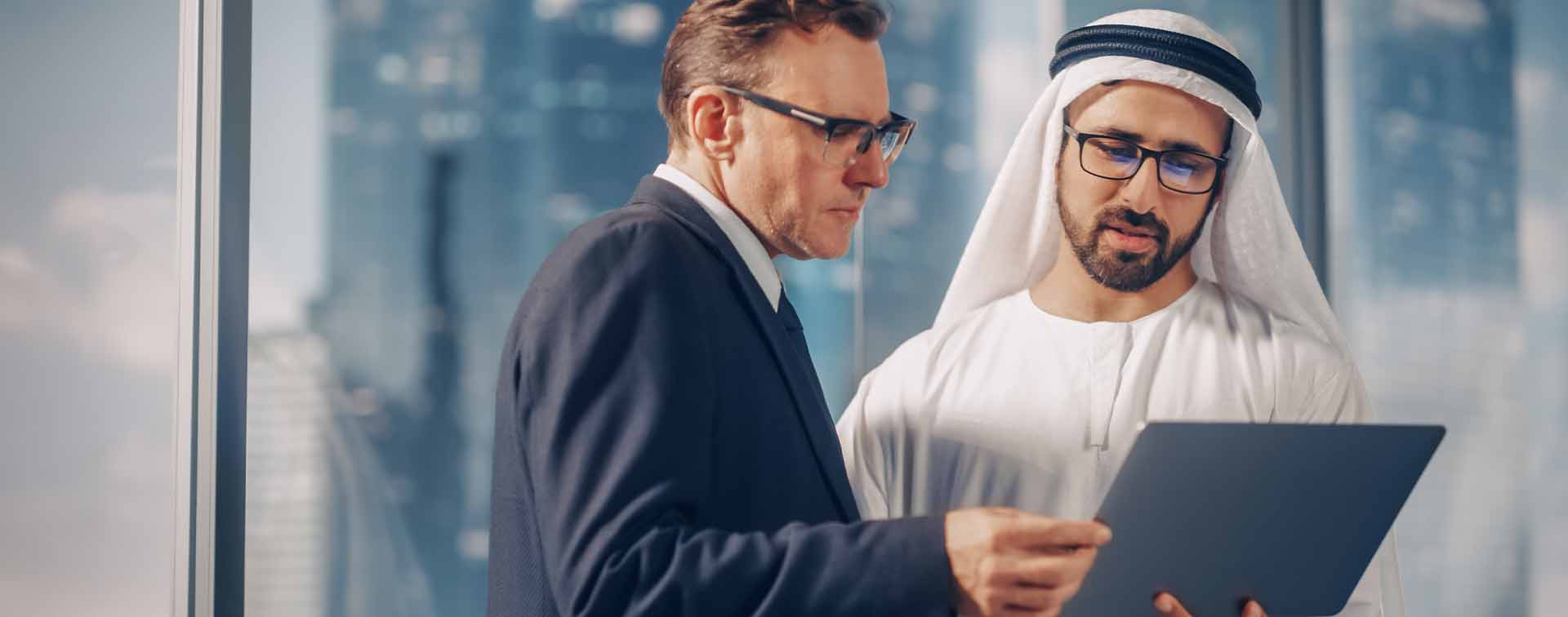 US businessman in suit meeting Emirati business partner in traditional kandura standing in modern office, looking at laptop