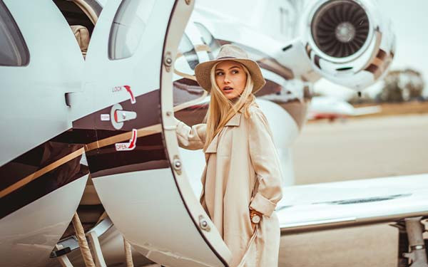 Young wealthy blonde female wearing beige hat and coat looking over her shoulder while embarking on a private airplane on a runway