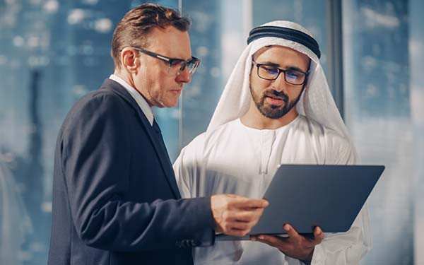 US businessman in suit meeting Emirati business partner in traditional kandura standing in modern office, looking at laptop
