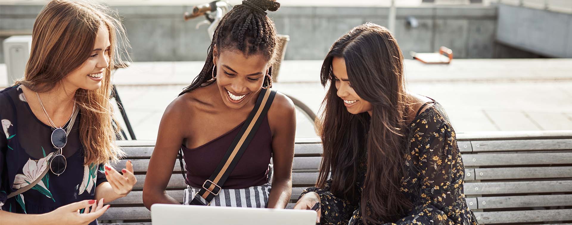 Three young female university students smiling while looking at a laptop on a bench outside